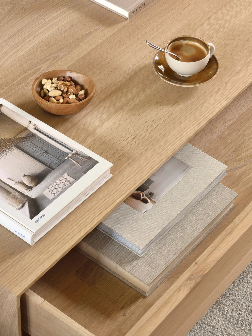 Ethnicraft Nordic Coffee Table, a close-up of the 120cm oak tabletop, highlighting its smooth, natural wood texture, with a coffee cup, bowl of nuts, and books elegantly arranged on its surface and inside the drawer.