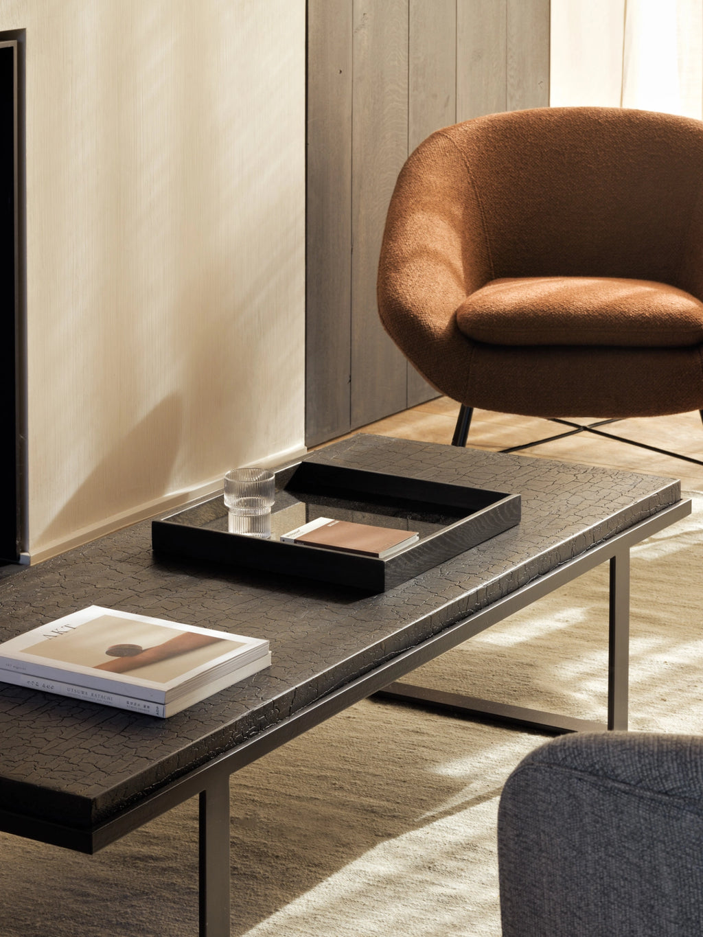 Ethnicraft Stability Coffee Table in umber, a rectangular modern coffee table with a richly textured, dark brown cracked finish, styled in a contemporary living room with a cozy orange armchair and light rug.