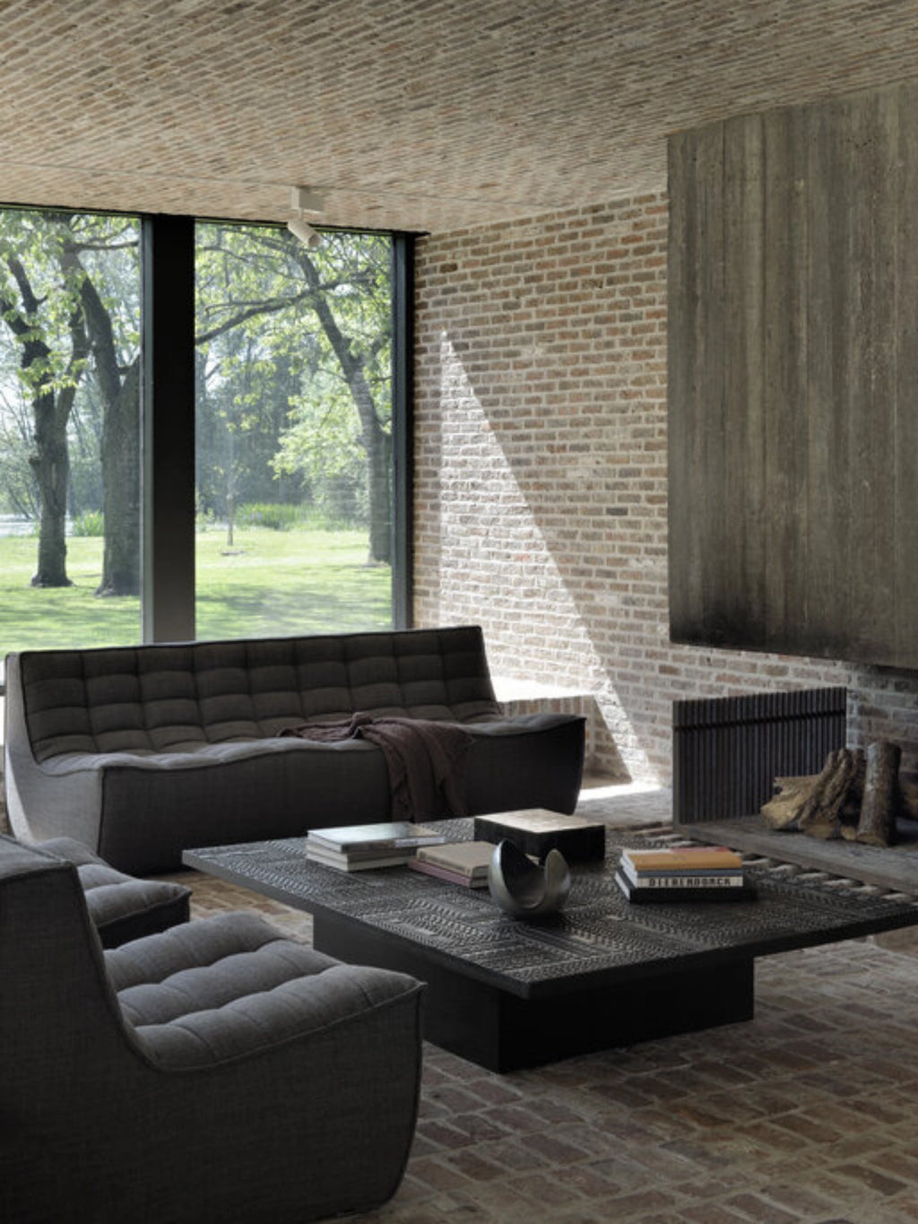 Ethnicraft Tabwa Coffee Table, a large rectangular black table with exquisite carved patterns, anchoring a stylish living room with a dark gray sofa and a rustic brick floor, exuding a modern industrial vibe.