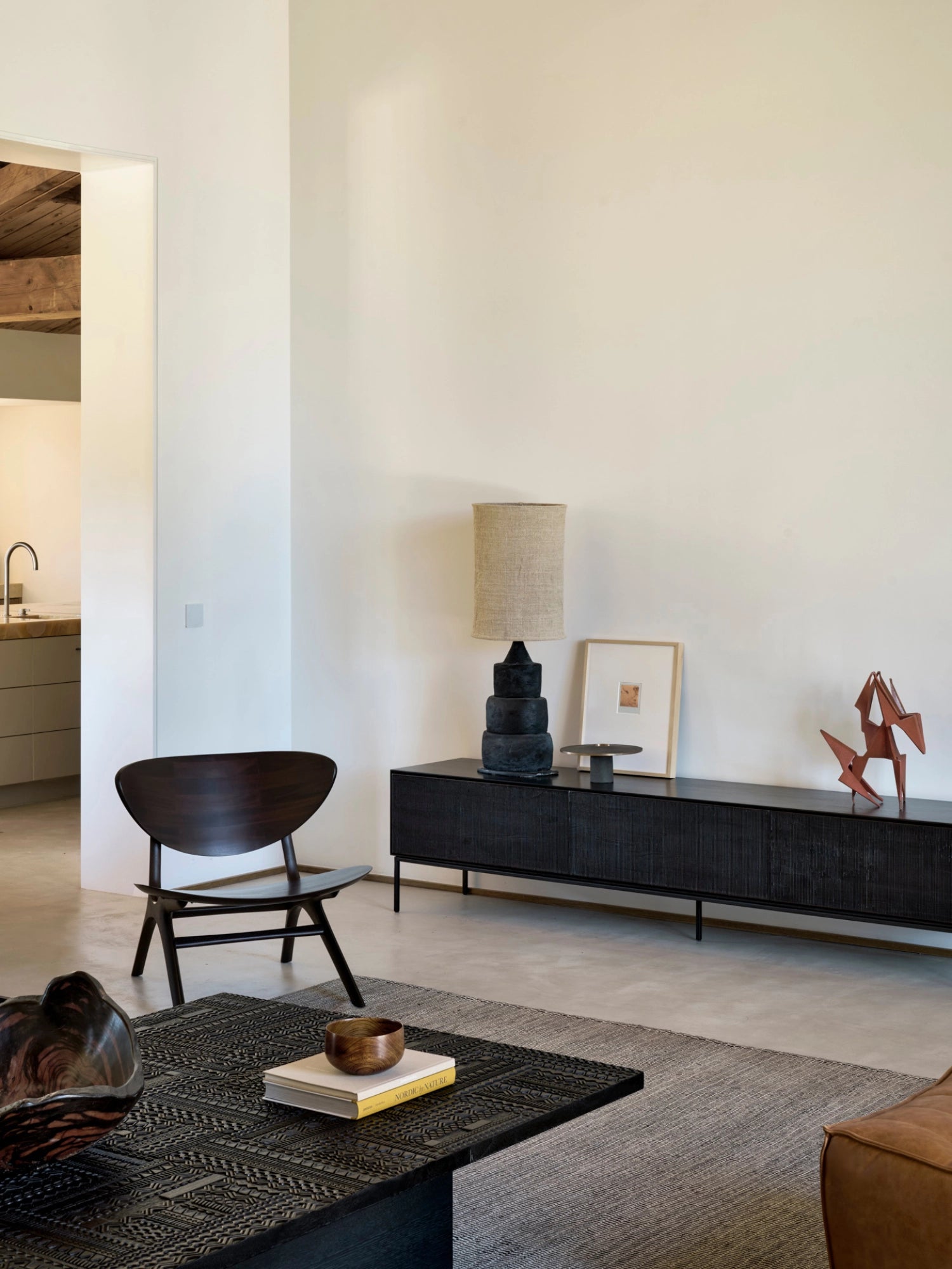 Ethnicraft Tabwa Coffee Table, a striking rectangular black table with intricate carved patterns, serving as a focal point in a modern living room with a dark wooden chair and minimalist decor.