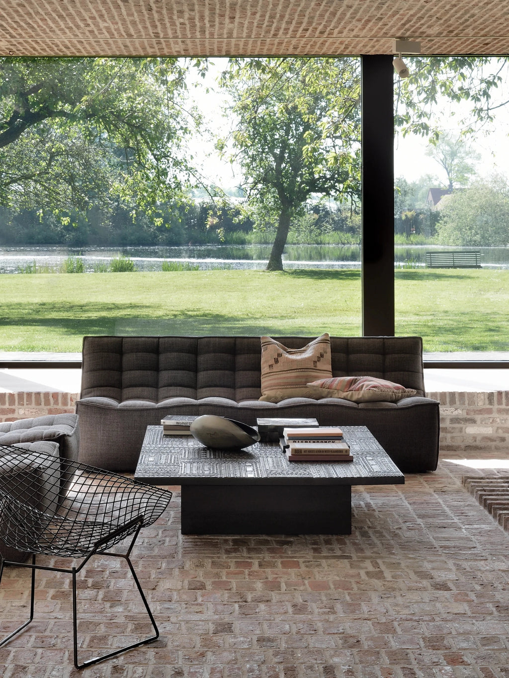 Ethnicraft Tabwa Coffee Table, a grand rectangular black table showcasing intricate carved designs, centrally placed in a spacious living area with a dark gray sofa and large windows overlooking lush greenery.