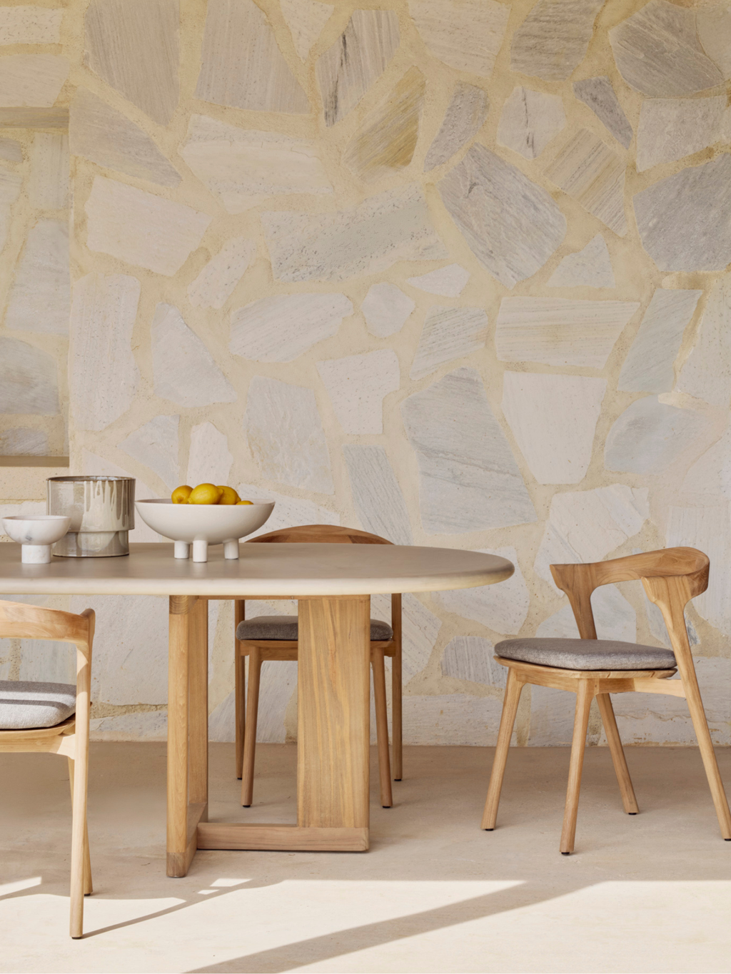 Ethnicraft Trestle Outdoor Dining Table, a contemporary oval outdoor dining table crafted from natural solid teak wood with a light grey composite tabletop and a distinctive rectangular trestle base. This organic modern teak dining set, complete with matching teak chairs featuring curved backs and light grey cushions, is ideal for stylish patio dining and sophisticated outdoor living spaces.