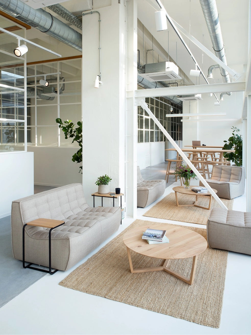 Ethnicraft Tripod Coffee Table in natural oak, featured in a bright, modern office lounge. The round, light wood table with its distinctive tripod base sits on a jute rug, surrounded by neutral-toned seating and green plants.