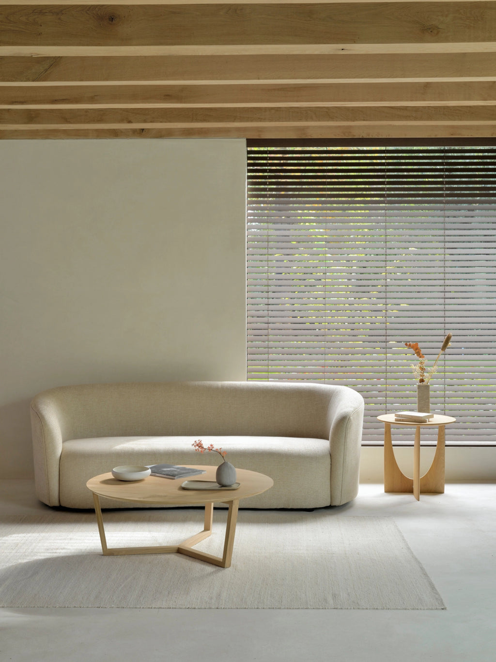 Ethnicraft Tripod Coffee Table in natural oak, positioned in a cozy, sunlit living room. The round, light wood table with its unique tripod base is placed on a light rug, complementing a cream sofa and a side table with decor.