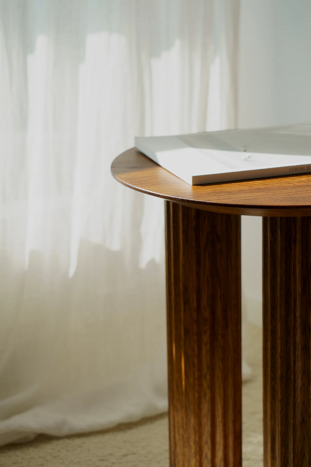 TAMO Filar Side Table, a premium round-top side table crafted from dark brown fluted wood, shown with a minimalist magazine on top, against a sheer white curtain. Perfect for modern living spaces.