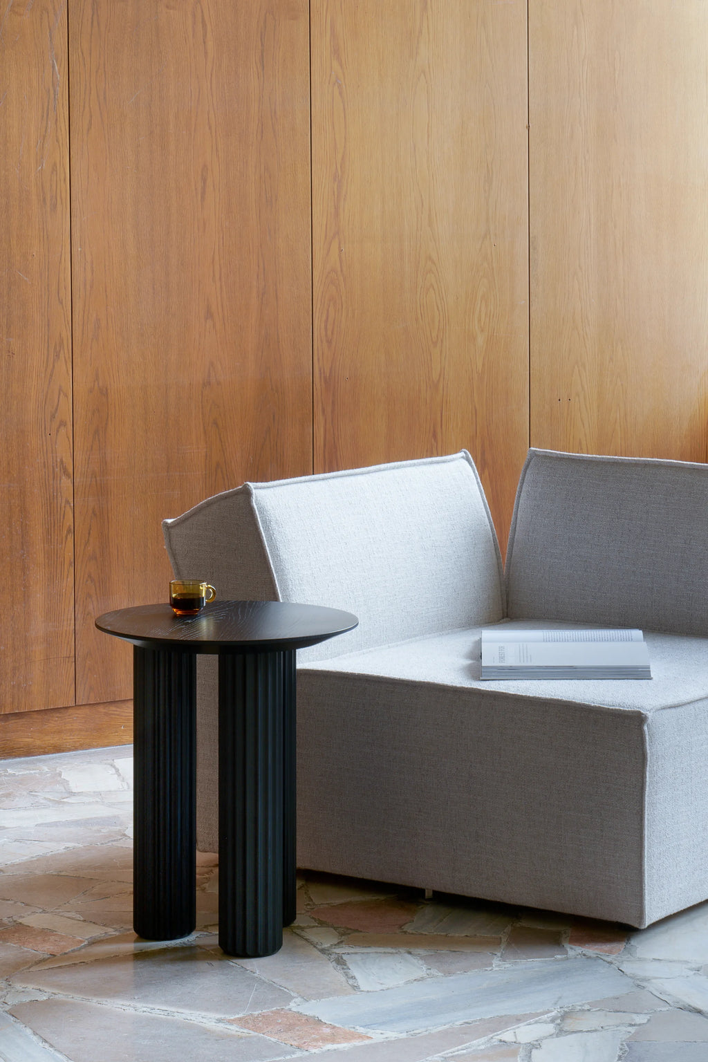 TAMO Filar Side Table, a sophisticated black column-leg table with a smooth round top shown with a golden cup, styled next to a light grey sofa. This luxury accent piece is ideal for modern living spaces.