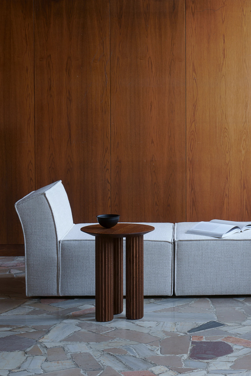 TAMO Filar Side Table, a premium dark brown solid wood table with a round top and three fluted legs, holding a black bowl, shown next to a beige sofa in a modern setting. Perfect for contemporary living spaces.