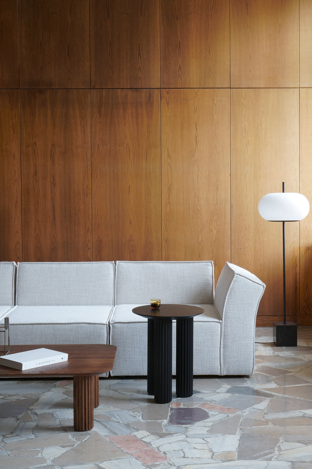 TAMO Filar Side Table, a modern black round-top table with three thick fluted legs, stylishly paired with a light grey couch. This premium accent piece is ideal for contemporary living spaces.