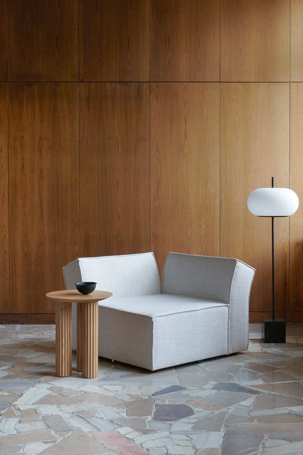 TAMO Filar Side Table, a light wood-colored round top table with fluted cylindrical legs, next to a light grey modern chair in a spacious living area. This authentic wooden accent table is perfect for contemporary decor.