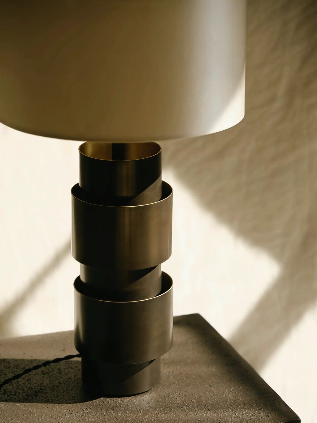 CTO Lighting Ring Table Lamp, close-up view showing the elegant bronze stacked cylindrical base and a light cream fabric shade, casting soft shadows on a textured surface.