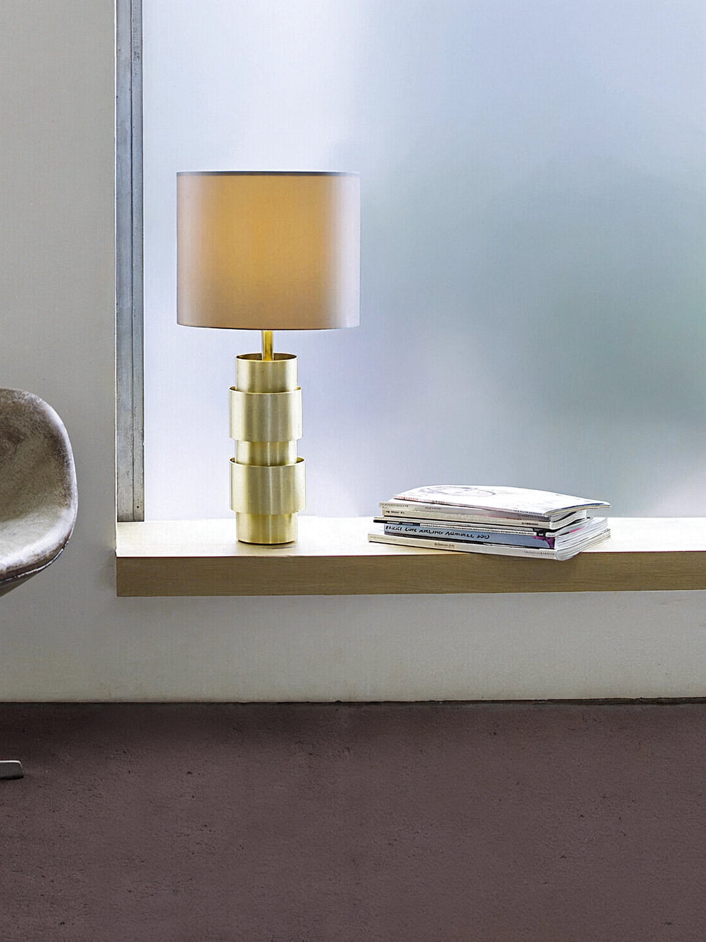 CTO Lighting Ring Table Lamp in a warm brass finish, with a tiered, sculptural base and a beige drum shade, placed on a light wooden window ledge next to stacked magazines.