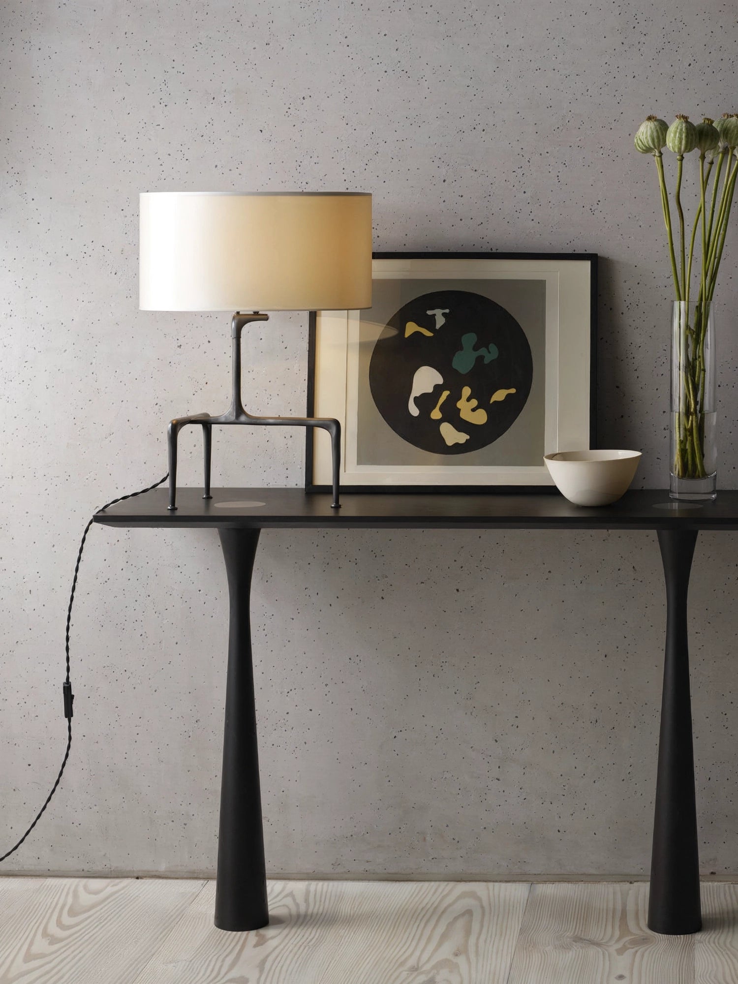 CTO Lighting Braque Table Lamp, a modern and elegant piece with a bronze-finished base and a cream fabric shade, casting a warm glow, displayed on a dark console table against a textured grey wall, beside a contemporary art print.