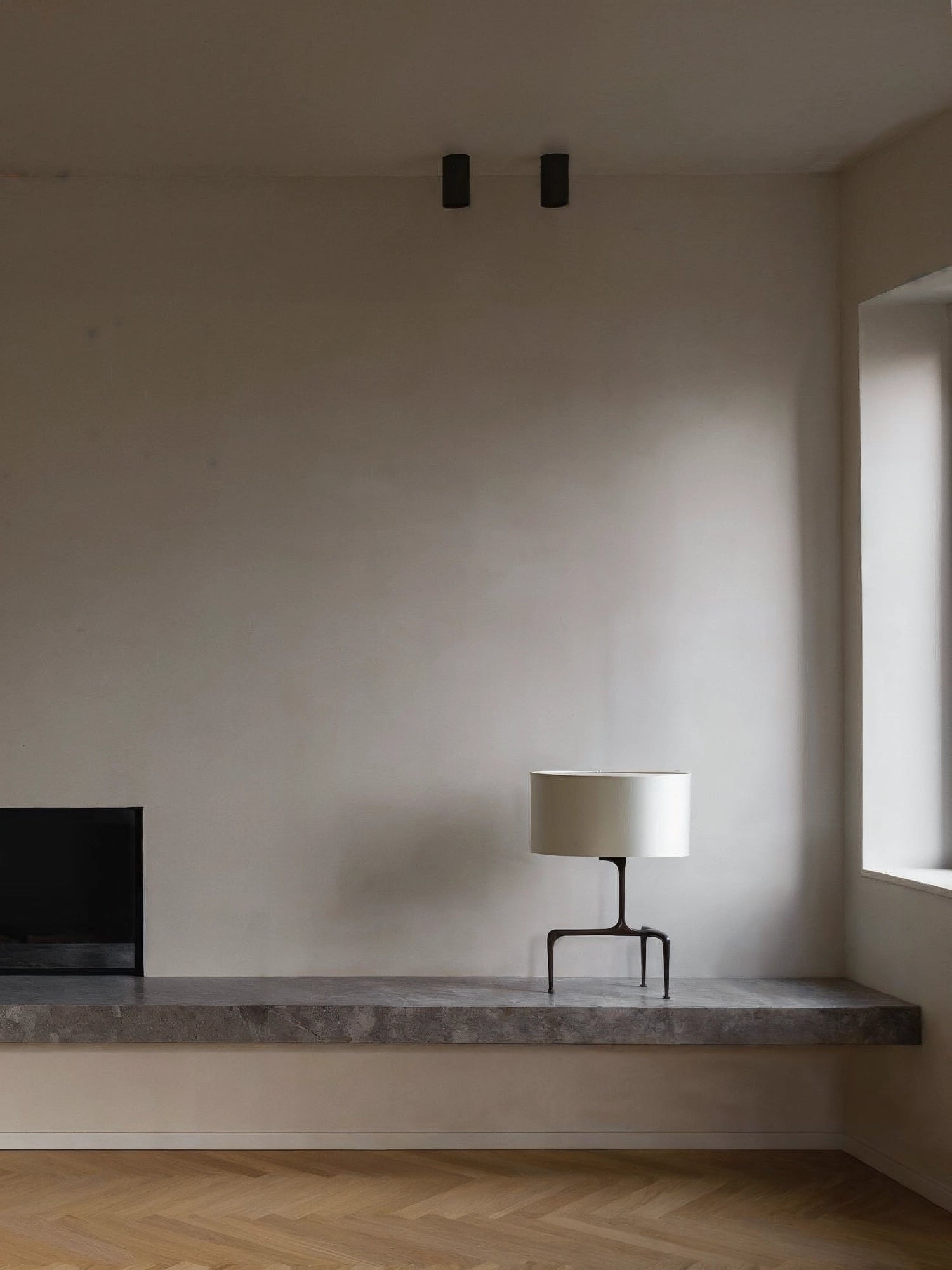 CTO Lighting Braque Table Lamp, a modern design with a light cream cylindrical shade and a dark bronze sculptural base, sitting on a concrete shelf in a minimalist room.