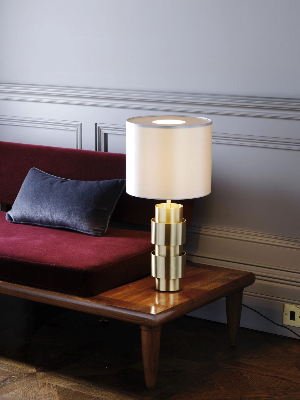 CTO Lighting Ring Table Lamp in a polished brass finish, featuring a multi-tiered cylindrical base and a bright white drum shade, sitting on a wooden side table next to a red sofa.