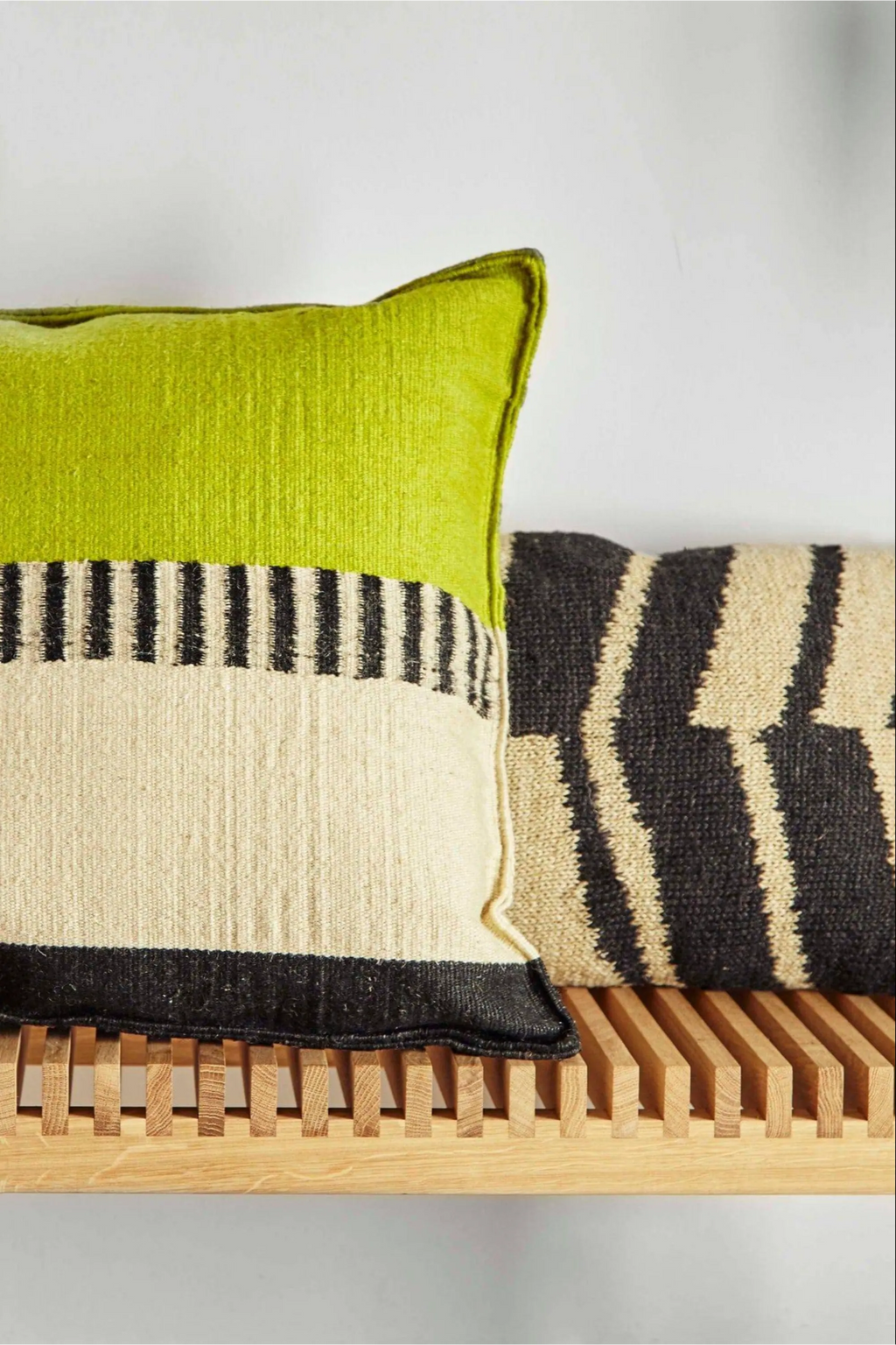 GAN Rugs Rustic Chic Cushion - Black & White, featuring a striking lime green top, natural beige body with black stripes, and a black and white geometric patterned cushion beside it, resting on a wooden slatted bench.