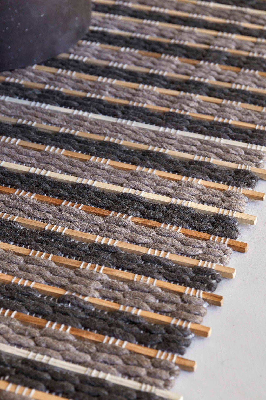 GAN Rugs Tsuchi Rug, a hand-woven organic modern area rug featuring alternating stripes of chunky natural wool in dark grey, light grey, and off-white, interwoven with natural wood slats. This textured, rustic design creates a unique visual and tactile experience, ideal for grounding minimalist living rooms, japandi bedrooms, or contemporary dining areas.
