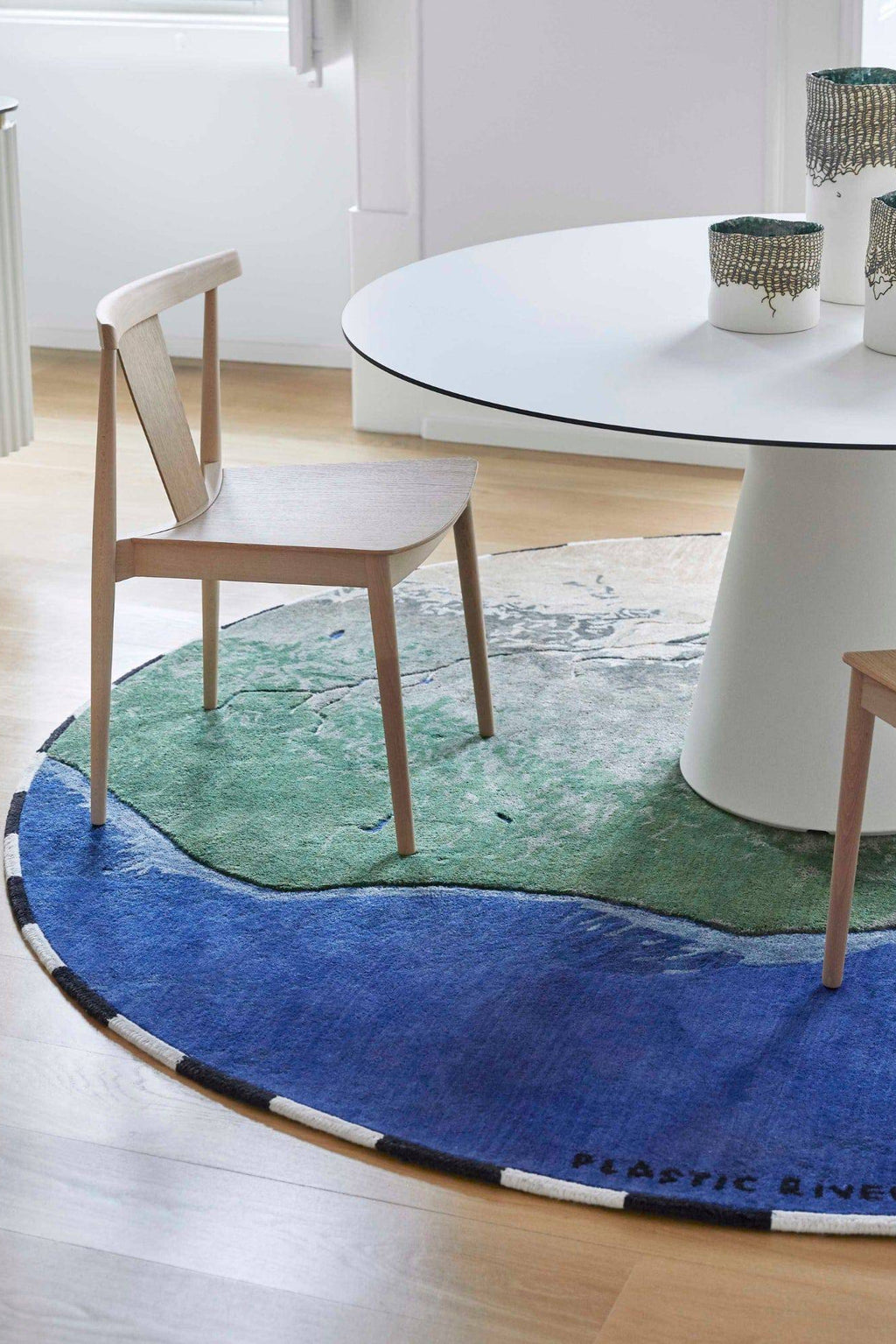 GAN Rugs Niger Rug, a striking round hand-tufted wool area rug featuring an abstract, nature-inspired design in vibrant royal blue, emerald green, and earthy tones, depicting a winding river landscape. This contemporary artistic rug has a plush texture, a contrasting black and white striped border, and subtly incorporates the 'PLASTIC RIVER' message, making it perfect as a statement piece for modern dining rooms, living rooms, or any eco-conscious interior seeking unique floor art.