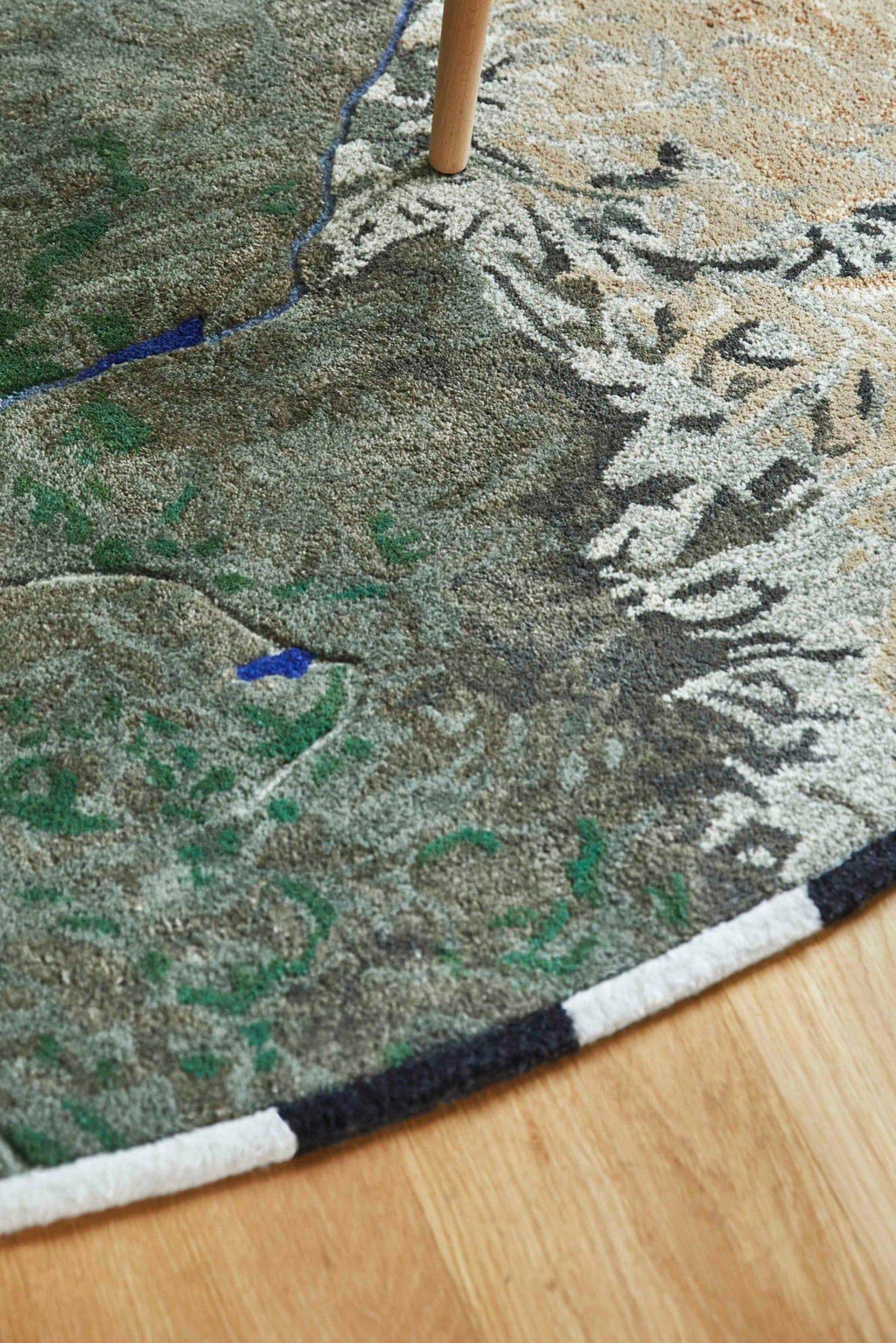 GAN Rugs Niger Rug, a hand-tufted wool area rug featuring an abstract, topographic landscape design in earthy tones of green, brown, beige, and grey. Deep blue accents mimic rivers and lakes, while varying pile heights create a textured, three-dimensional effect. A distinct black and white striped border adds a graphic touch. This organic modern wool rug is perfect for adding a nature-inspired accent to contemporary living rooms, bedrooms, or dining areas.