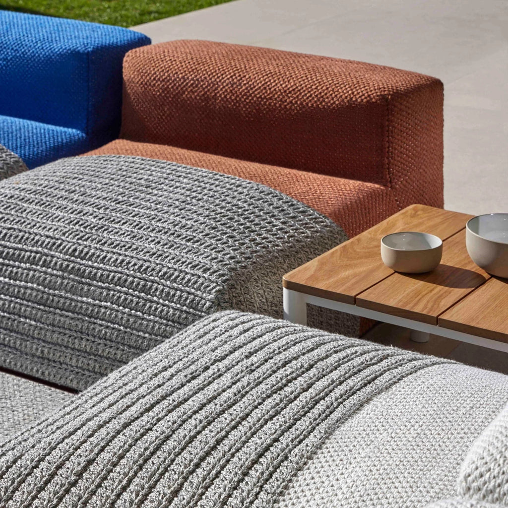 GAN Rugs Mangas Outdoor Composition 2, a modern modular outdoor sofa featuring vibrant blue and burnt orange tightly woven fabric backrests, complemented by light and dark grey textured knit cushions with distinct ribbed patterns. This weather-resistant outdoor lounge furniture set includes a contemporary side table with a natural wood slatted top and a clean white metal frame. Ideal for creating a stylish and comfortable outdoor living space, perfect for patios, decks, or garden lounge areas.