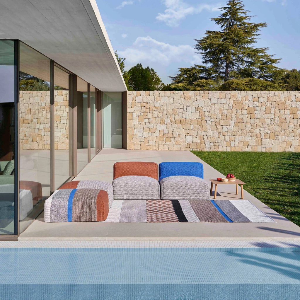 GAN Rugs Mangas Outdoor Composition 2, a vibrant modular outdoor sofa and matching rug set featuring chunky hand-knitted fabric in a multi-colored striped pattern of terracotta, royal blue, grey,.