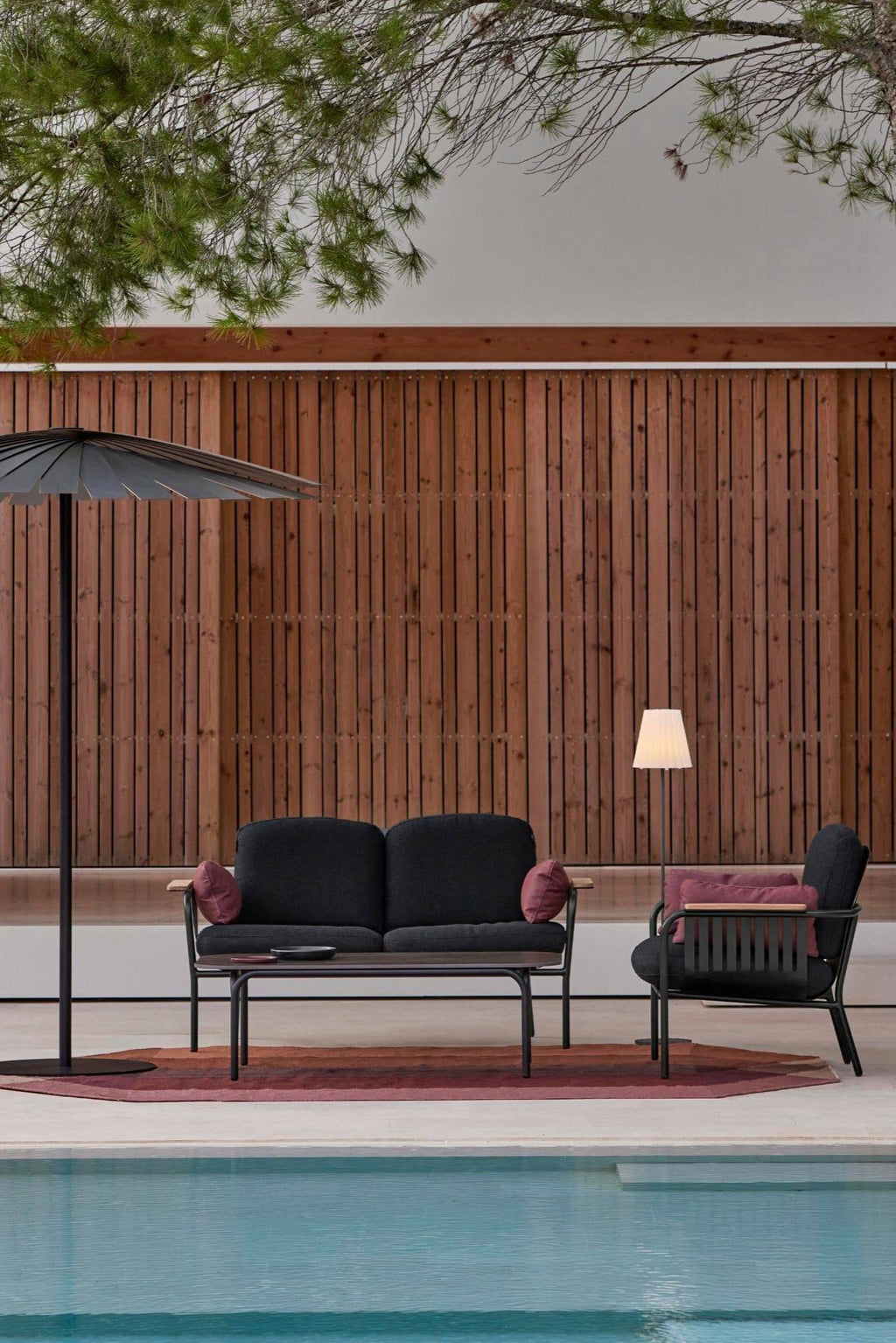 Gandia Blasco Capa 2-Seat Sofa, a modern minimalist outdoor sofa featuring a sleek matte black aluminum frame and plush dark gray upholstered cushions, complemented by integrated natural wood side tables on the armrests. This contemporary 2-seater outdoor lounge is perfect for luxury patio seating, poolside relaxation, or a minimalist garden space.