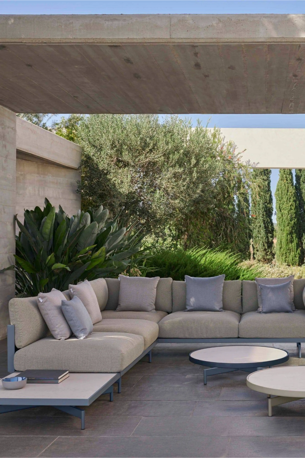 Gandia Blasco Onde Sectional 1, a modern minimalist L-shaped outdoor sofa featuring light beige weather-resistant fabric upholstery and a sleek matte grey powder-coated aluminum frame. This contemporary modular lounge includes coordinating light grey and pale blue accent cushions, offering stylish comfort and clean lines. Ideal for luxury outdoor living spaces, modern patios, and sophisticated poolside relaxation areas.