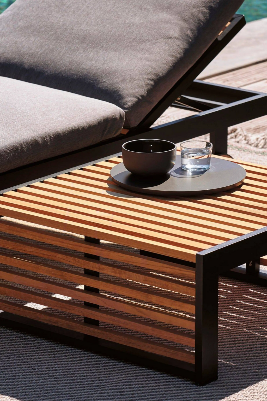 Gandia Blasco DNA Teak Side Table, a sleek modern outdoor table featuring natural warm teak wood slats for the top and sides, supported by a durable matte dark grey powder-coated aluminum frame with clean lines. This minimalist slatted design patio table is perfect for contemporary outdoor living spaces, poolside lounges, or garden seating areas.