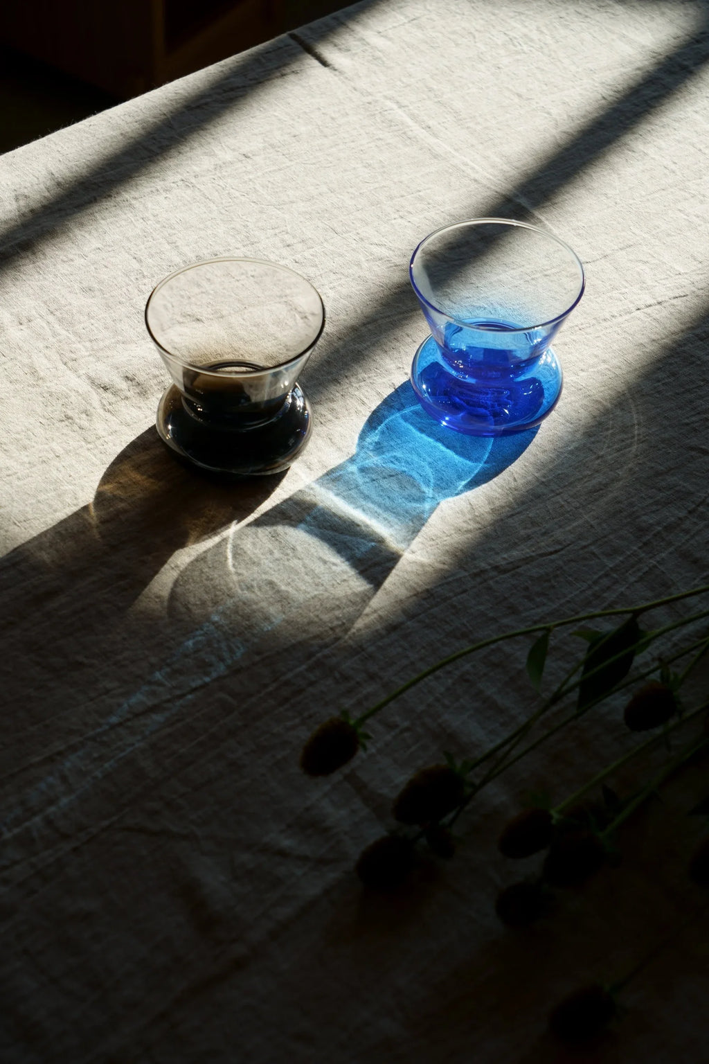 TAMO Orio Drinking Glass - Set of 2: One smoky transparent gray, one vibrant blue, hourglass-shaped glasses displayed on a textured beige linen fabric with high-contrast sunlight and shadows. These unique drinking glasses are perfect for elevating everyday beverages.