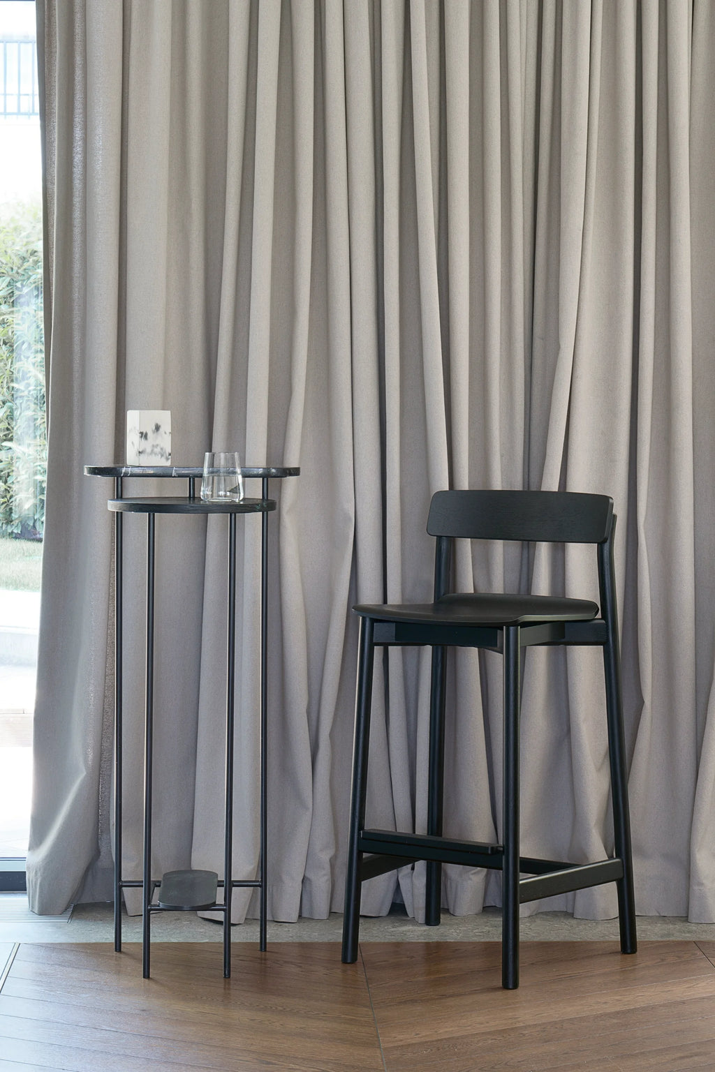 TAMO Arche Bar Stool, a sleek black wooden high chair with a backrest and footrest, displayed on a wooden floor next to a tall black metal table. Offers premium, minimalist design. Ideal for modern kitchen islands or bar areas.