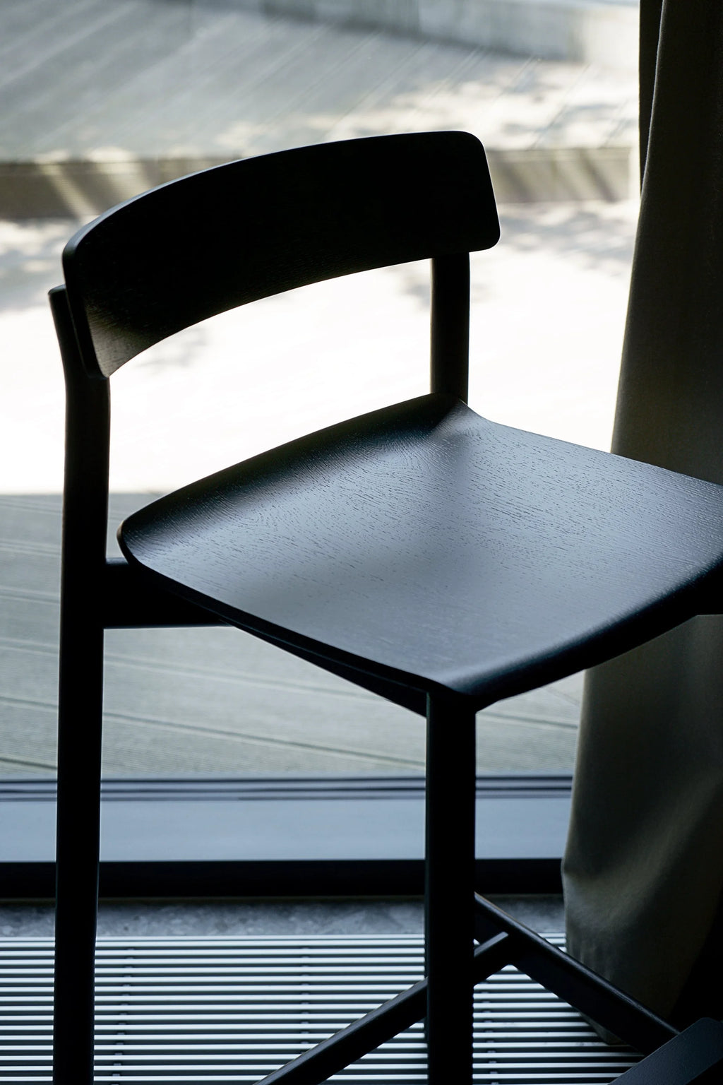 TAMO Arche Bar Stool, a sleek black modern bar stool with a textured matte wooden seat and backrest, displayed indoors by a window with natural light. A premium contemporary design piece.