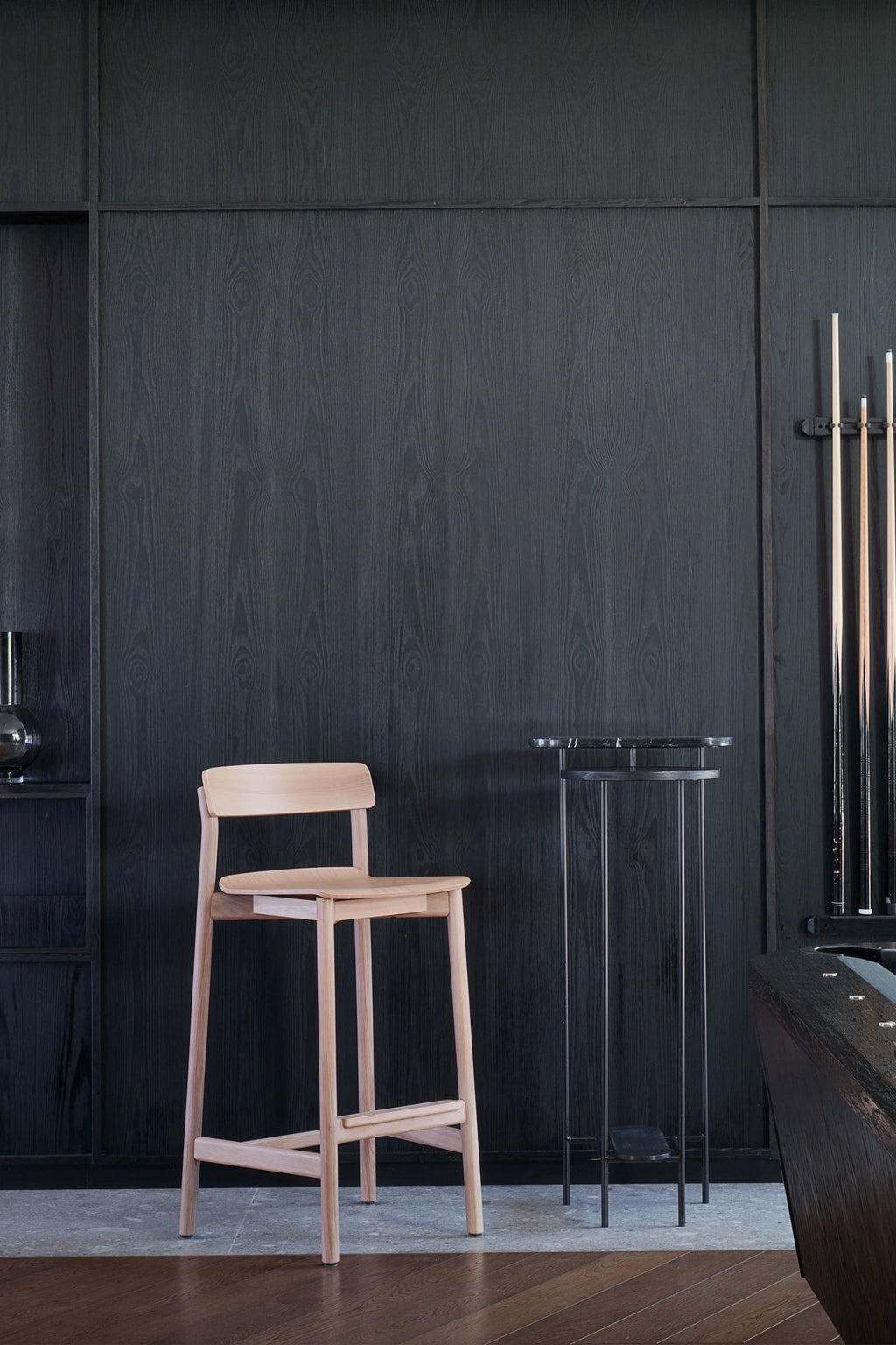 TAMO Arche Bar Stool, a premium light beechwood bar stool with a minimalist design, small backrest, and footrests, displayed in a dark wood-paneled room. Ideal for modern kitchens or bars.