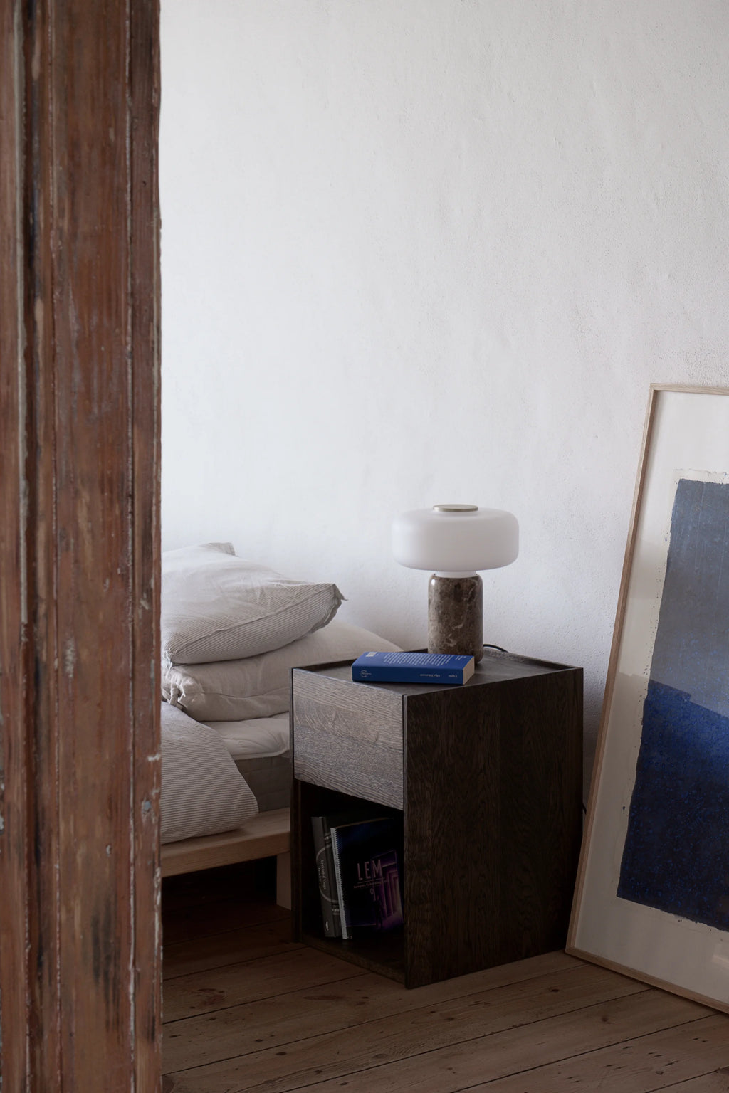 TAMO Kari Nightstand, a modern dark oak wood nightstand with a subtle grain texture, open shelf, and drawer, styled with a contemporary lamp and book on top in a bedroom setting. Ideal for stylish bedside storage.