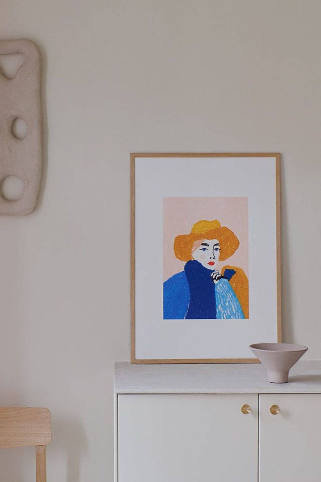 The Poster Club Woz, a vibrant modern portrait art print featuring a woman with a wide-brimmed yellow hat and blue coat, rendered in a painterly, textured illustration style with a soft pink background and striking red lips. This colorful contemporary wall art is framed in a light natural oak frame with a white mat, enhancing its artistic appeal. Ideal as a statement piece for modern living rooms, creative home offices, or a dynamic addition to a gallery wall.