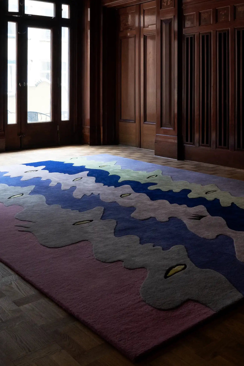 Layered Opera Goers Wool Rug, a unique abstract area rug featuring wavy patterns in shades of blue, purple, gray, and pink, with subtle yellow accents. Perfect for adding a modern, artistic touch t...