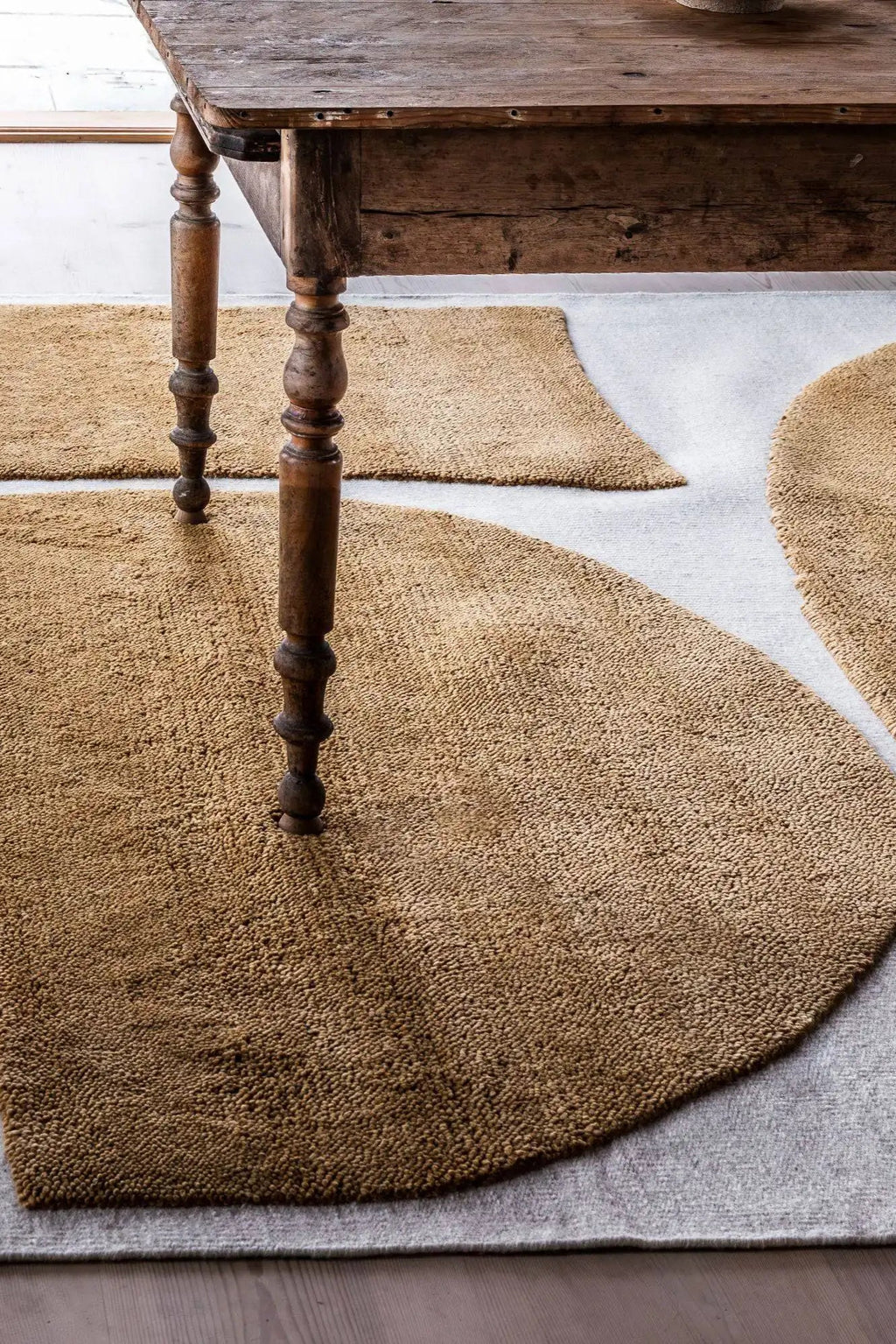 Layered Gotland Rauk Ullmatta Wool Rug, a thick pile, naturally textured brown wool rug with an organic, rounded design, peeking out from under a rustic wooden table. Perfect for adding warmth and...