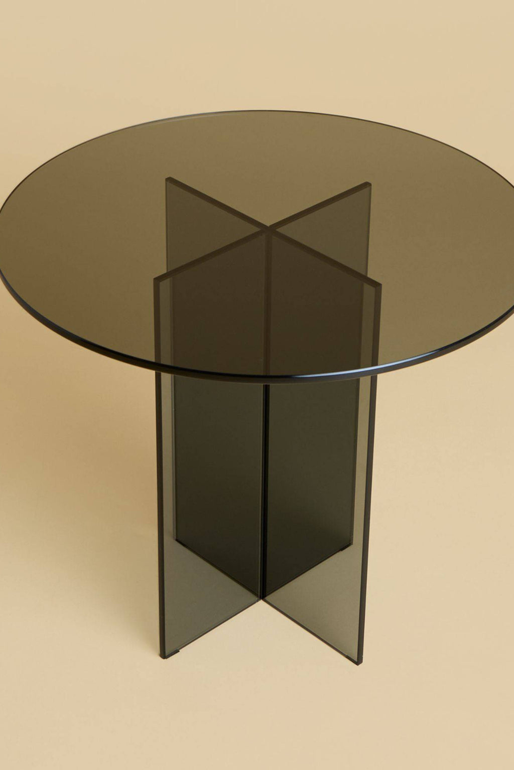 Lebanto Amami Side Table, a modern minimalist side table crafted from dark smoked glass with a sleek round tabletop and a striking geometric cross-shaped pedestal base. This contemporary glass accent table adds a sophisticated touch to any living room, bedroom, or entryway with its clean lines and reflective surface.