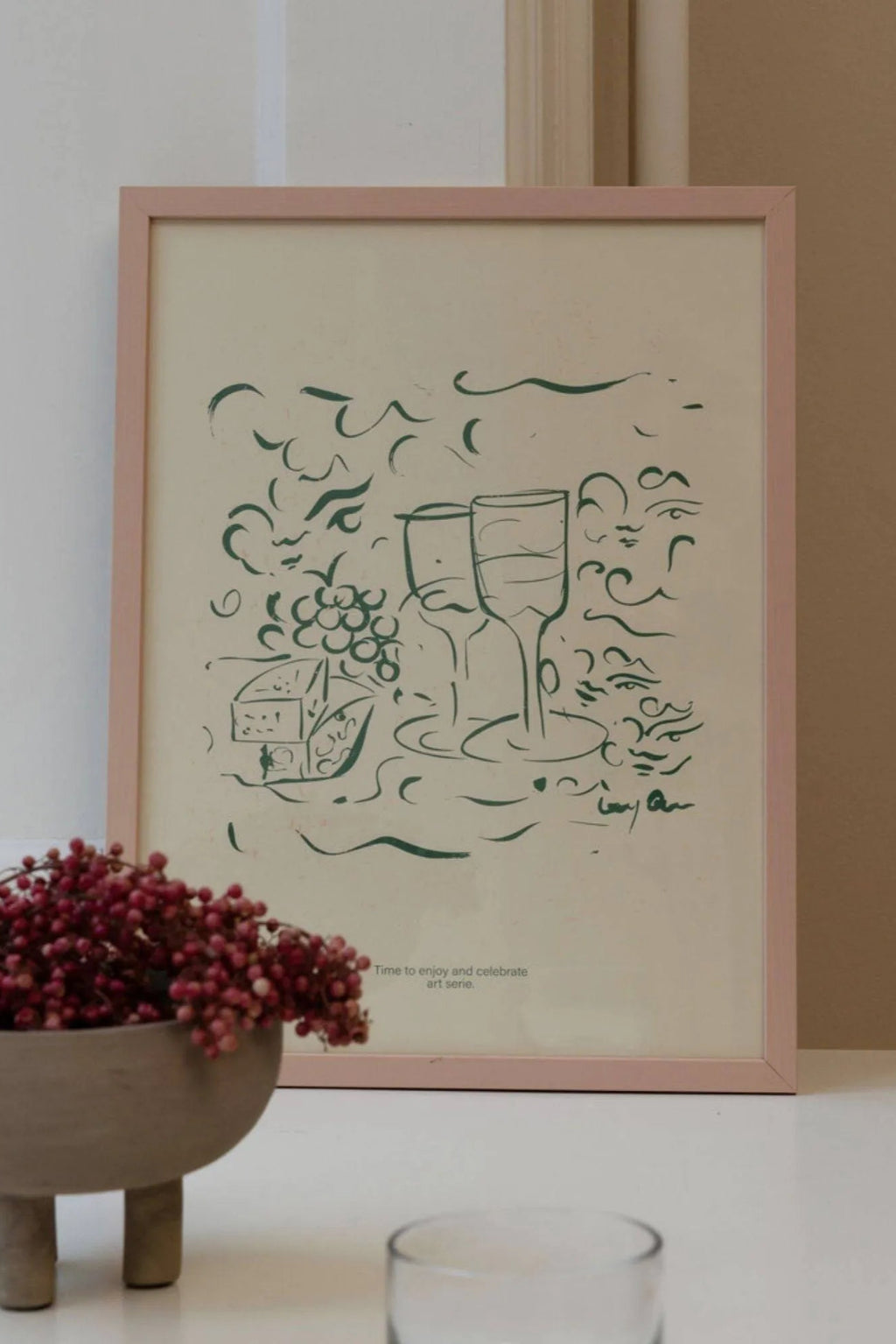 The Poster Club Cheese and Wine, a minimalist abstract art print featuring dark green line art illustrations of wine glasses, grapes, and cheese on a light beige background, housed in a thin light pink wooden frame. This contemporary wall art is perfect for kitchen decor, dining room styling, or a chic bar cart accent.