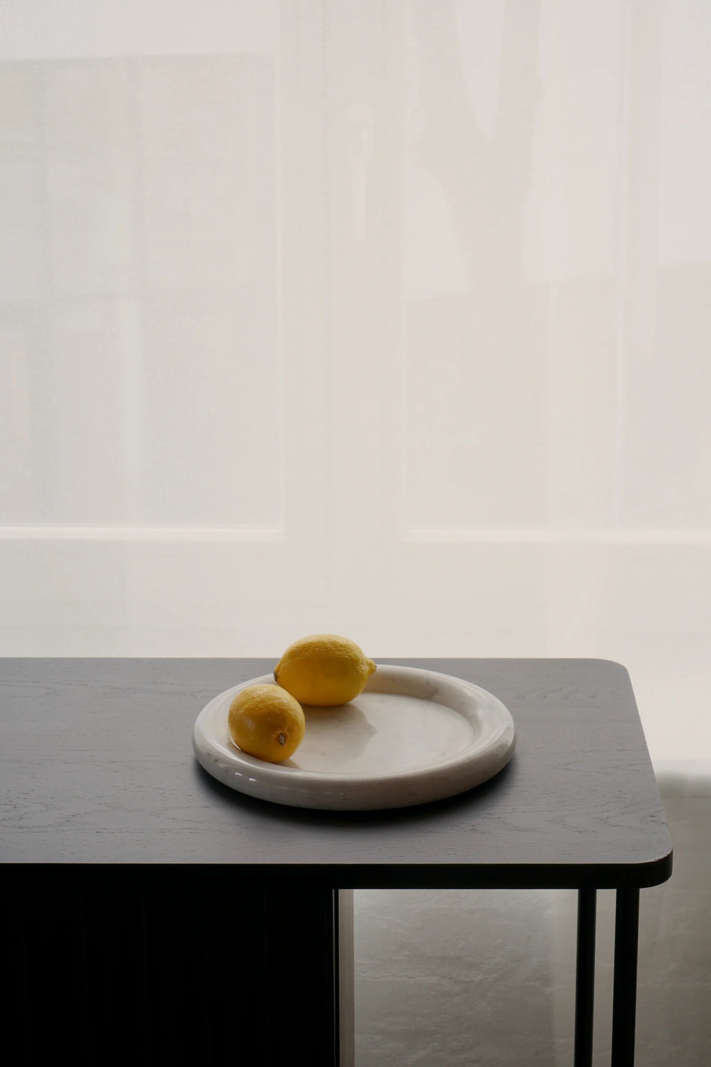 TAMO Luva Marble Tray shown in a lifestyle setting, a pale white exquisite marble round tray with two whole lemons, resting on a dark wood table. Ideal for kitchen or decorative use.