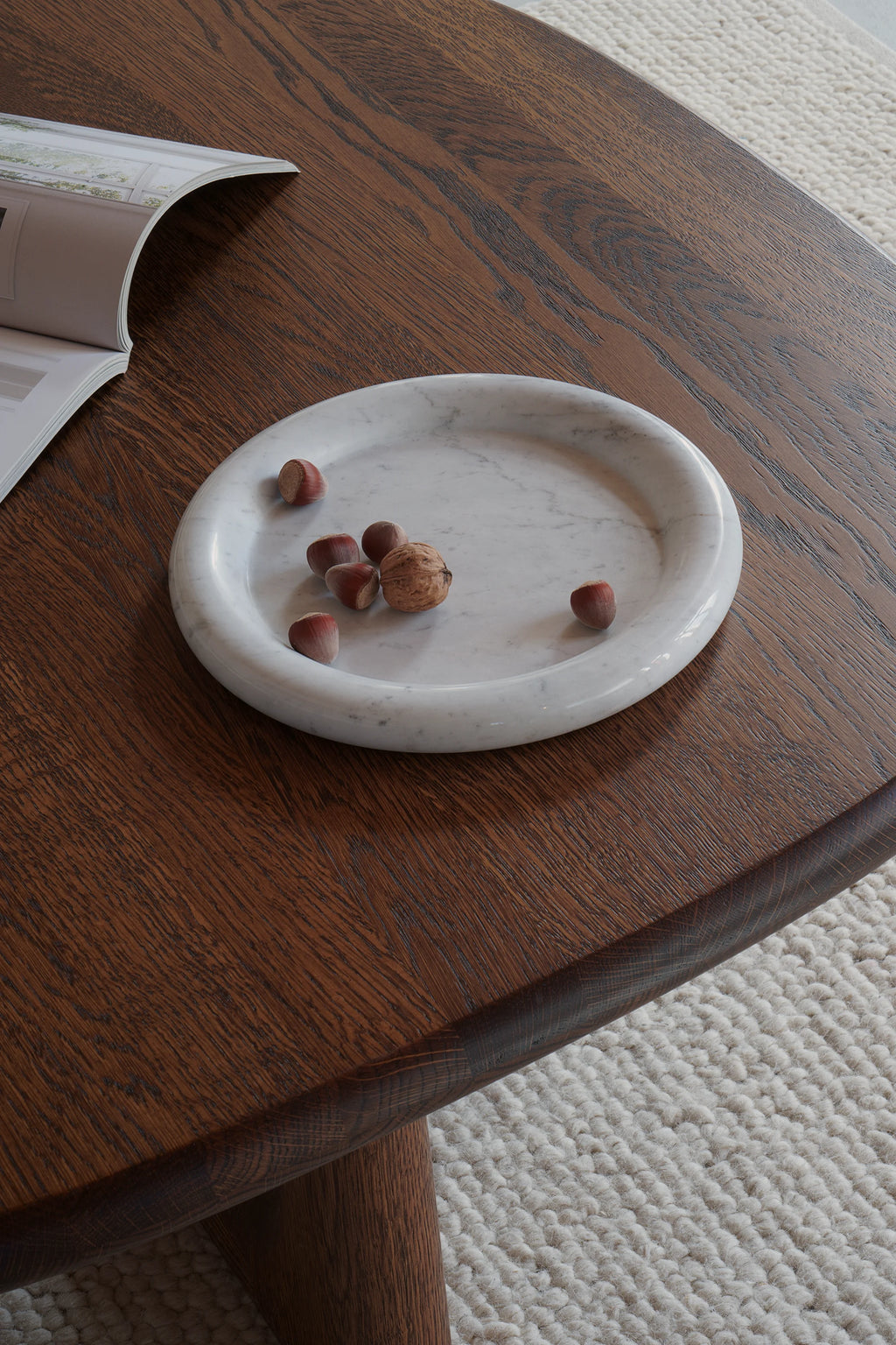 TAMO Luva Marble Tray: Round white marble tray with subtle grey veining and a raised, organic-shaped rim, displaying hazelnuts and a walnut on a rich brown wooden table. Perfect for elegant home decor.