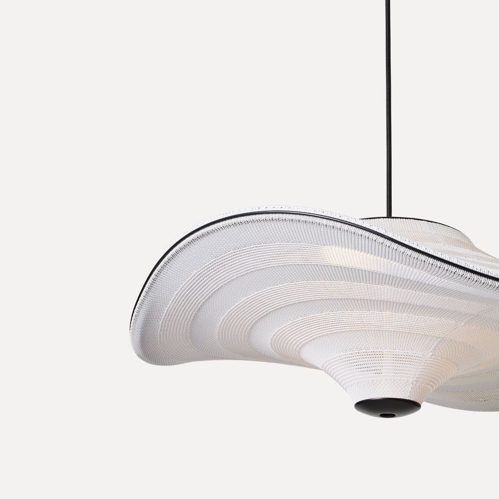 Made by Hand Flying Ø58 Pendant Lamp, a white organic modern pendant lamp featuring a fluid, sculptural shape crafted from finely woven textile with subtle ribbed detailing. Its distinctive undulating form is accented by a crisp black trim along the edge and a matte black base, creating a striking contrast. This contemporary ceiling light is ideal for minimalist living rooms, modern dining spaces, or as a statement piece in a sophisticated bedroom.