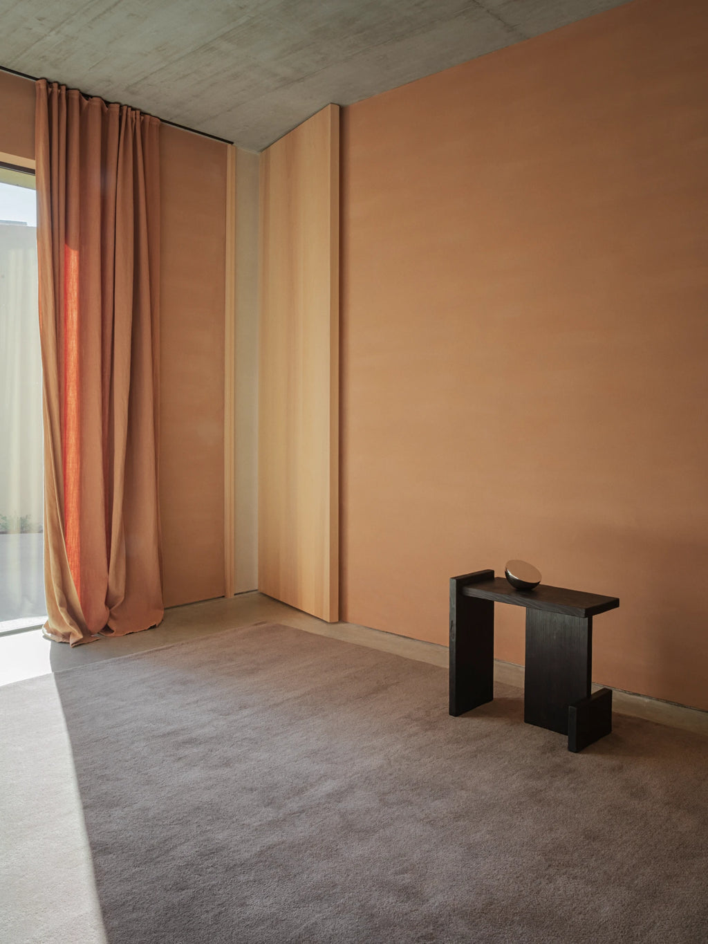 A spacious room with minimalist decor features a large, plush, hand-tufted rug in a deep gray-brown color. The walls are a warm terracotta shade, and the curtains are a similar earthy tone. A unique black side table with a square top and two legs sits on the rug. The room has a concrete ceiling and light wood panels near the window.
