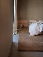 A cozy bedroom features a large, plush, hand-tufted rug in a neutral brown color. The rug is placed under a bed with a wooden headboard and a white comforter. A small, light-colored side table with a lamp is on the rug. The walls are a warm, earthy tone, and a long beige curtain hangs next to a window. The floor is a smooth concrete.