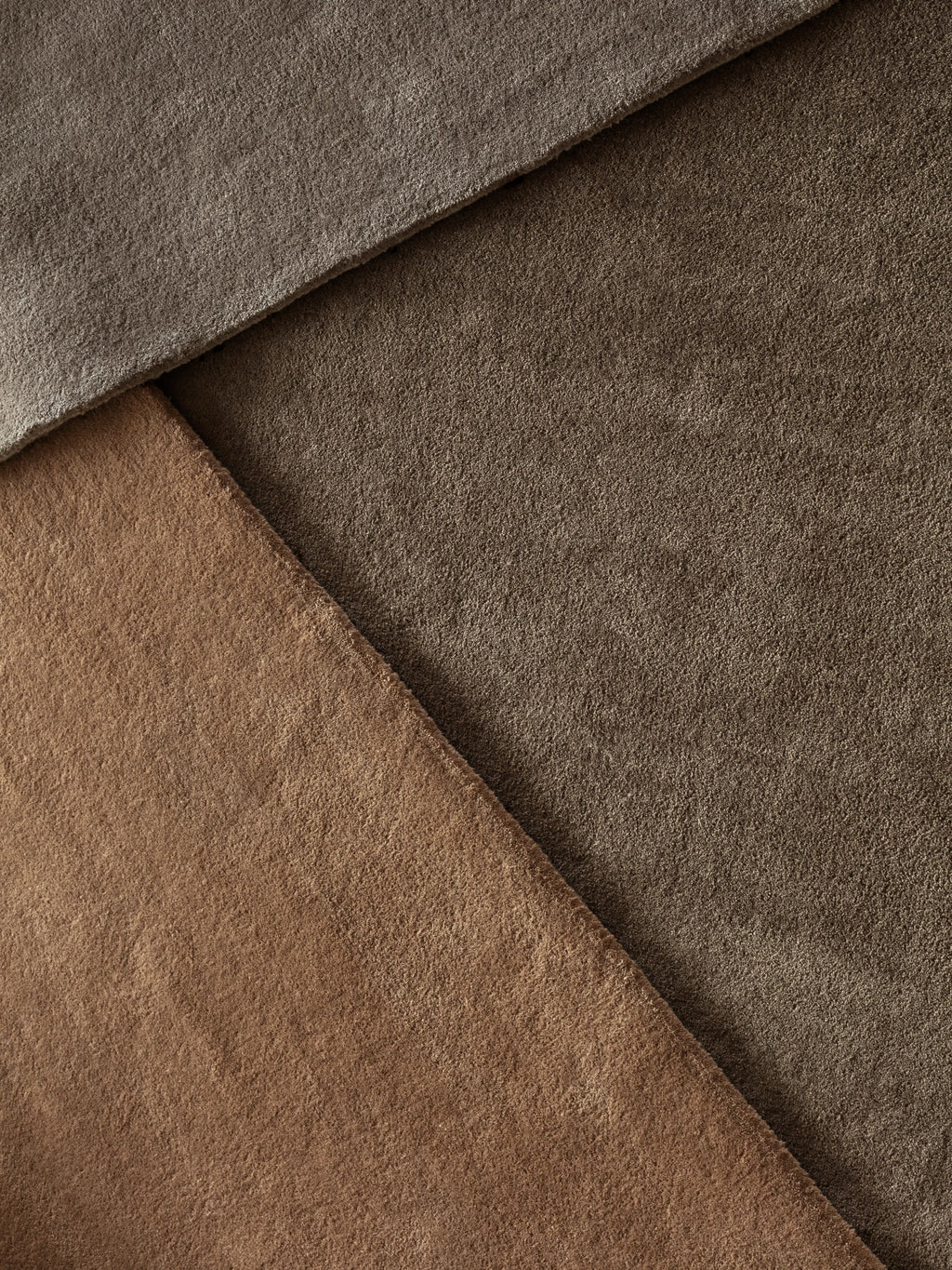 A close-up flat lay shows three overlapping swatches of plush, hand-tufted wool rugs. The swatches are in various earthy tones: a deep gray, a dark brown, and a lighter reddish-brown or clay color. The different textures of the rugs are visible, and the overall composition creates a diagonal pattern.
