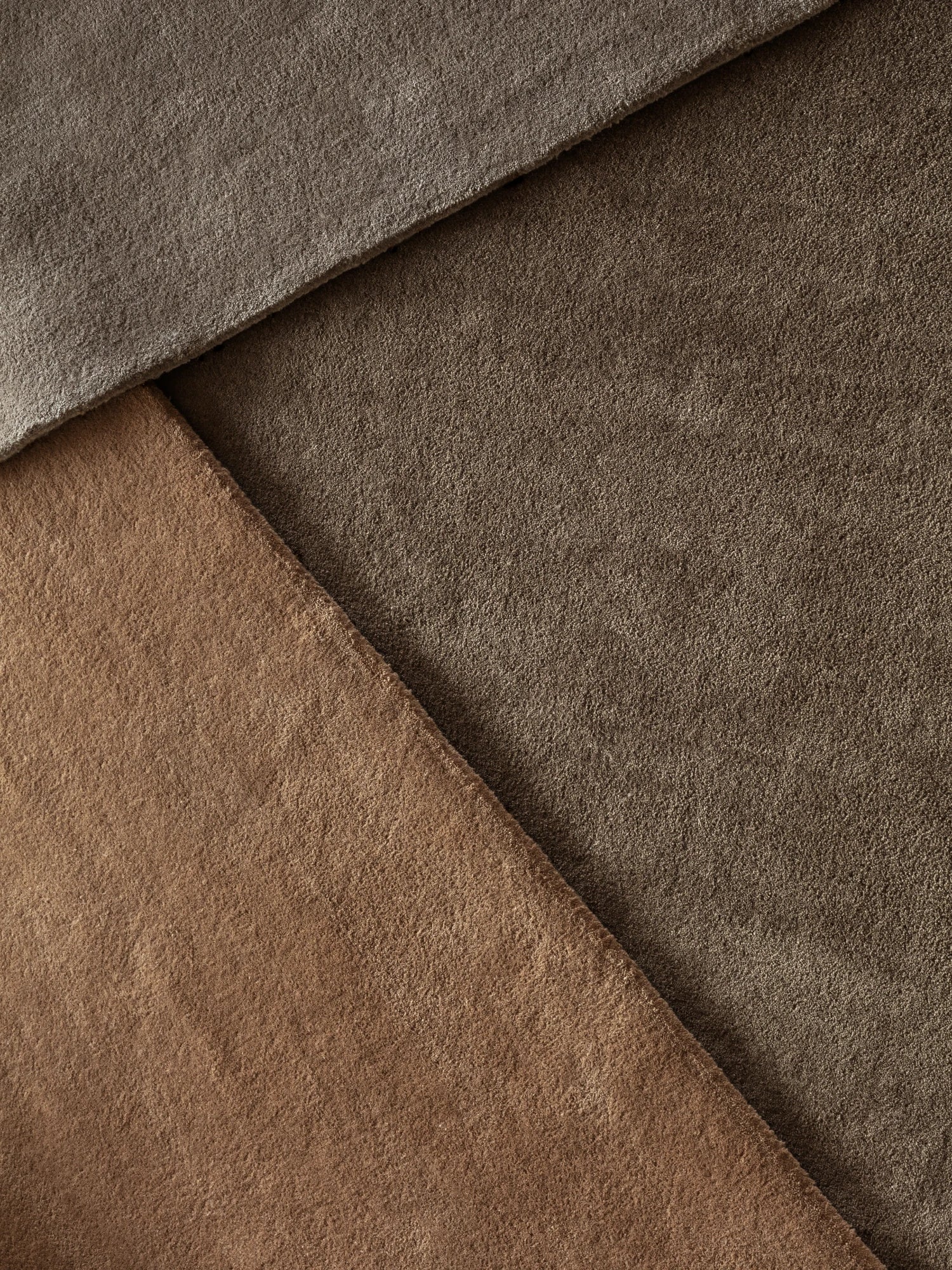 A close-up flat lay shows three overlapping swatches of plush, hand-tufted wool rugs. The swatches are in various earthy tones: a deep gray, a dark brown, and a lighter reddish-brown or clay color. The different textures of the rugs are visible, and the overall composition creates a diagonal pattern.