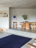 A long, plush area rug in a deep navy or marine blue color is on a gray concrete floor. The rug is placed in front of a raised dining area with a rustic wooden table with two thick, cylindrical terracotta-colored bases. A chair and a small stool are on either side of the table, and a blue and white striped vase with green leaves is on top of the table. A white wall with a framed blue circular art print and a wooden chair is behind the table.