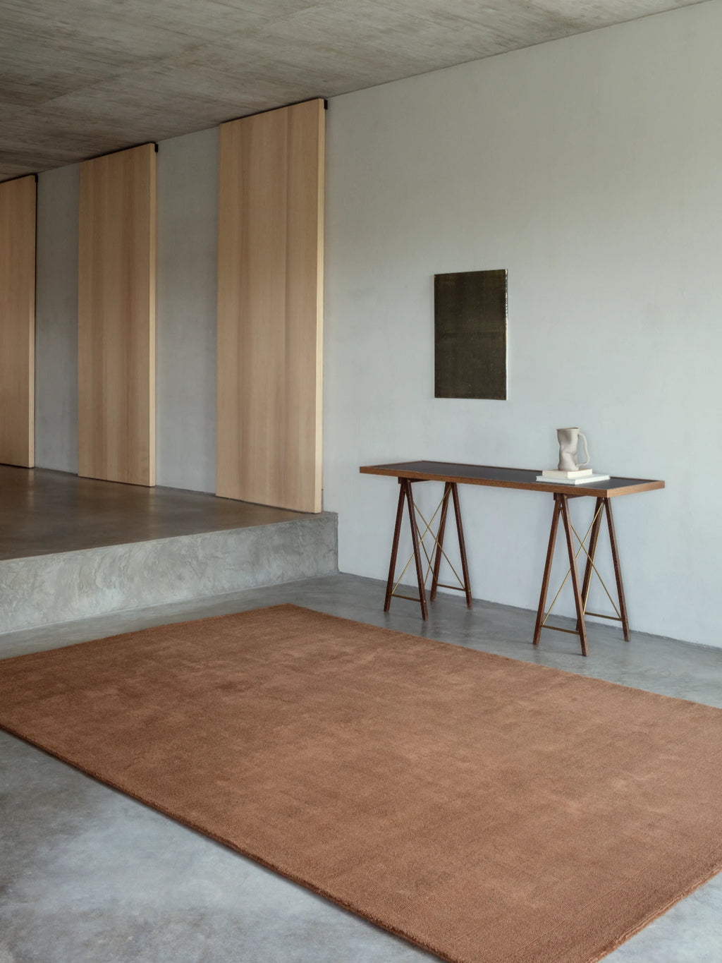 A large, plush area rug in a reddish-brown color is on a gray concrete floor. A modern console table with dark wooden trestle legs and a black top is against the wall, holding a small decorative vase and a stack of books. The room has a concrete ceiling, and light wood panels are on the wall to the left.