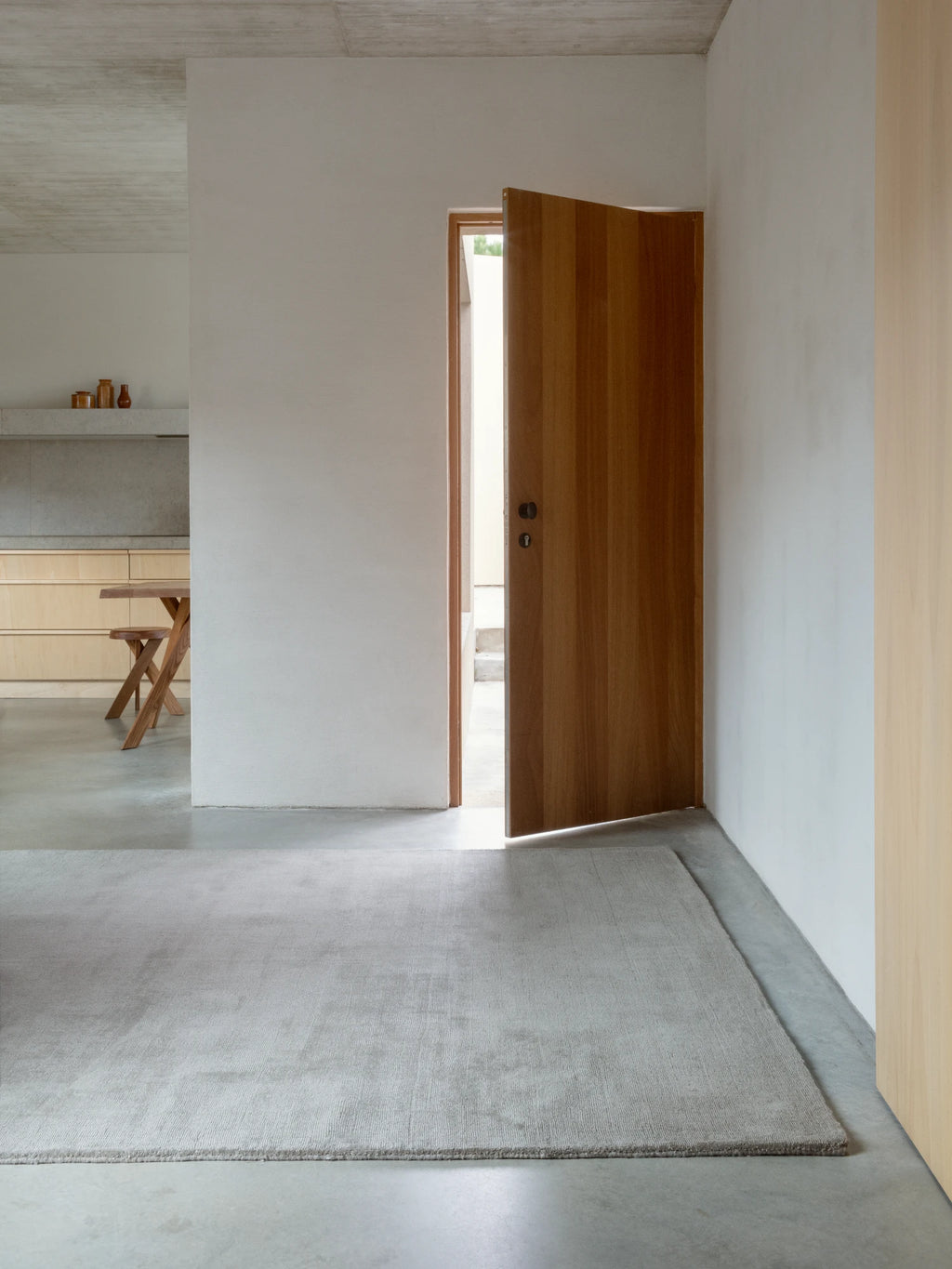 A long, rectangular wool rug in a light gray or beige color is on a concrete floor in a minimalist entryway. The rug has a subtle textured pattern. A wooden door is ajar in the background, leading to another room, and a partial view of a light wood kitchen or dining area is visible to the left.