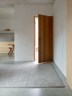A long, rectangular wool rug in a light gray or beige color is on a concrete floor in a minimalist entryway. The rug has a subtle textured pattern. A wooden door is ajar in the background, leading to another room, and a partial view of a light wood kitchen or dining area is visible to the left.