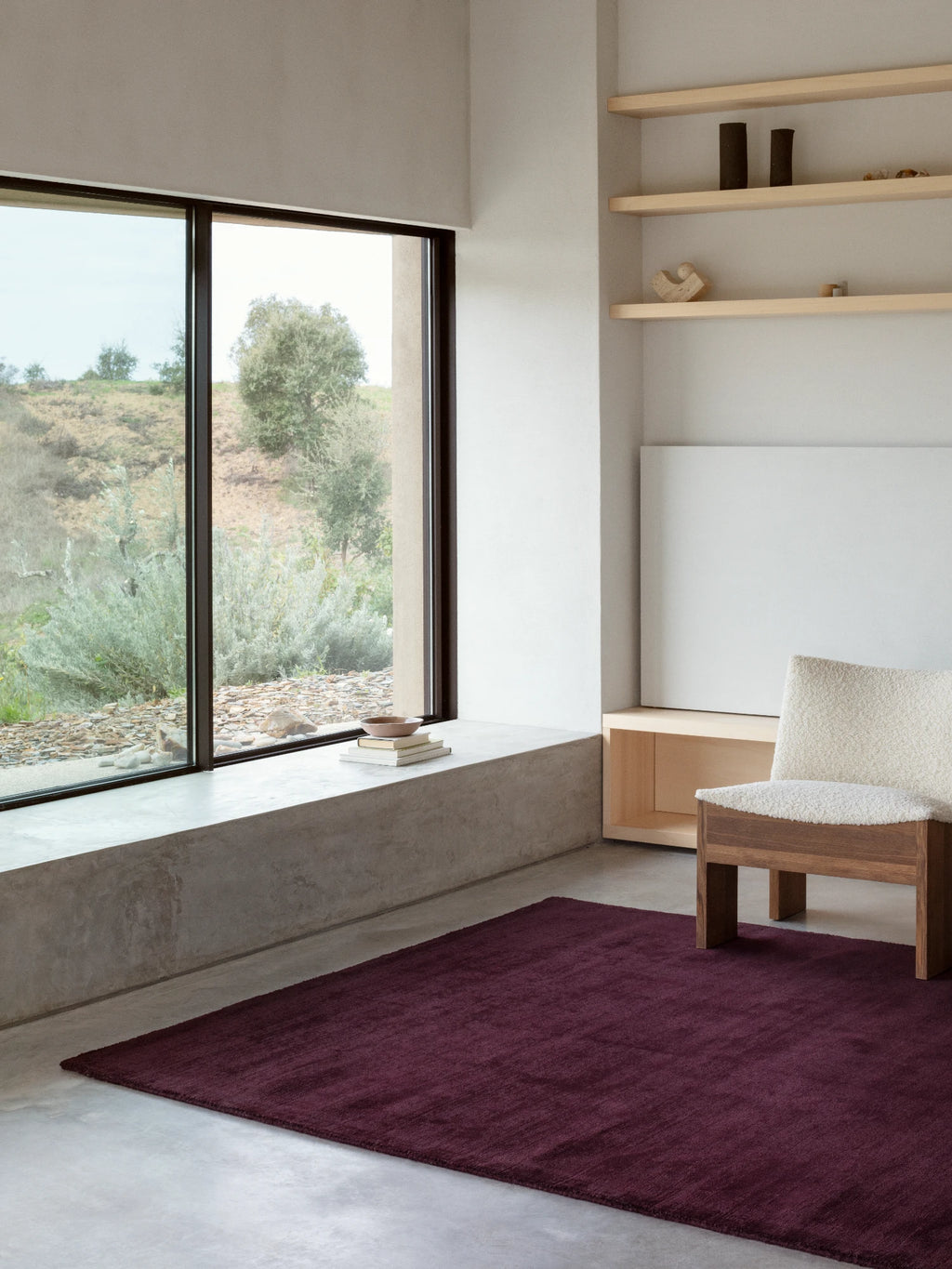 A large, rectangular area rug in a deep burgundy or dark wine color is on a gray concrete floor. The rug is placed in front of a wide window that looks out onto a hilly landscape with green and silvery-gray bushes. A light-colored chair with a fuzzy texture is on the rug, and a wall with built-in wooden shelves and a blank canvas is visible on the right.