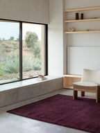 A large, rectangular area rug in a deep burgundy or dark wine color is on a gray concrete floor. The rug is placed in front of a wide window that looks out onto a hilly landscape with green and silvery-gray bushes. A light-colored chair with a fuzzy texture is on the rug, and a wall with built-in wooden shelves and a blank canvas is visible on the right.